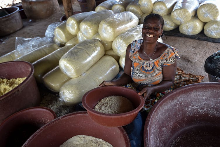 A smiling woman holds up a bowl of ground cassava. Bowls and storage bags of the food item surround her.