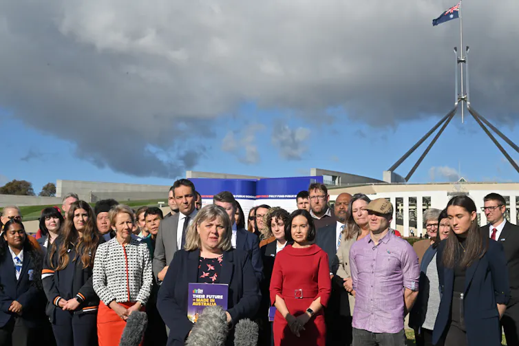 A group of adults, including state education ministers, stand on the front laws of Parliament House in Canberra. There are grey clouds in the sky above.