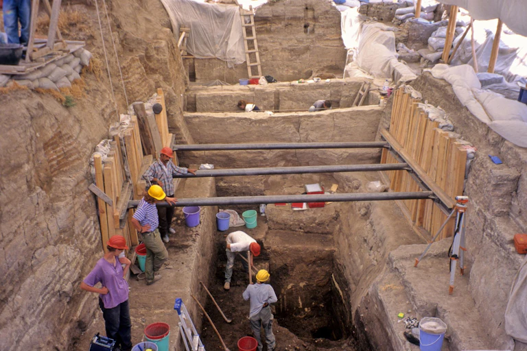 people in hardhats stand along perimeter of a trench in dusty land, a couple dig within