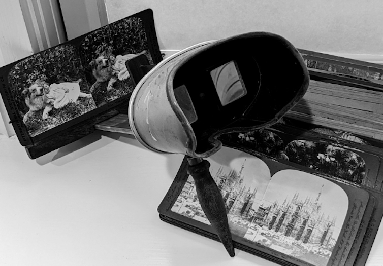 A metal stereoscope with a wooden handle and a card mounted in its frame is propped on a stack of other image cards.