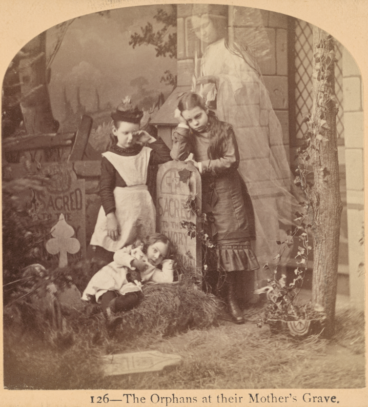 Three young girls with sad expressions lean on a gravestone; the ghost of a woman stands protectively behind them