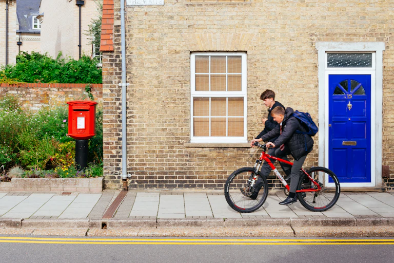 View from the street of two teen boys walking and cycling from school in front of a house
