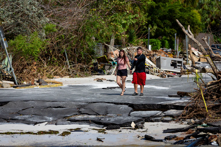 A woman and a man wearing shorts and t shirts hold hands and walk on a badly damaged road, with fallen trees and debris nearby them.