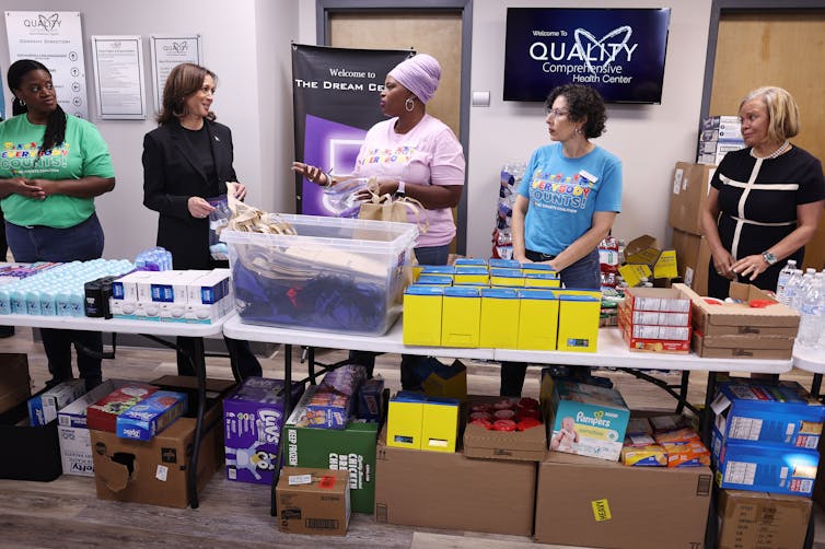Five women stand in line and look at each other, as they stand in front of a table that has boxes on it and under it.