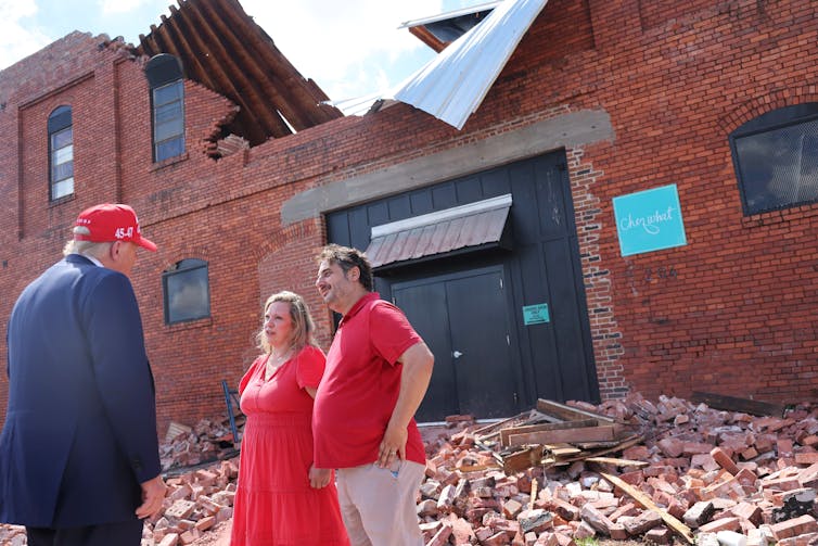 A man with a blue suit and red hat faces a man and a women wearing red and they stand in front of a partially destroyed red brick building with rubble on the ground.
