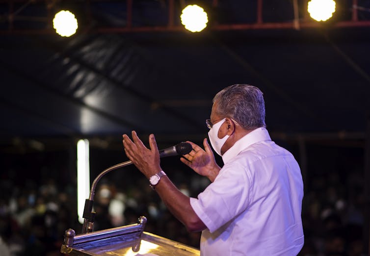 An Indian man wearing a face mask and a short-sleeved dress shirt faces mostly away from the camera while speaking to a crowd from behind a podium