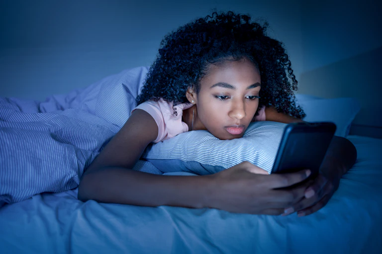 A teenaged girl lies on a bed using a smartphone
