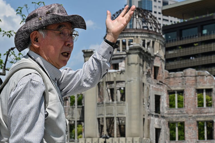 An elderly man gestures in front of a bomb-damaged building