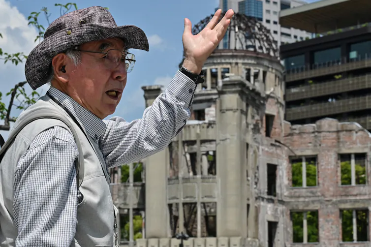 An elderly man gestures in front of a bomb-damaged building