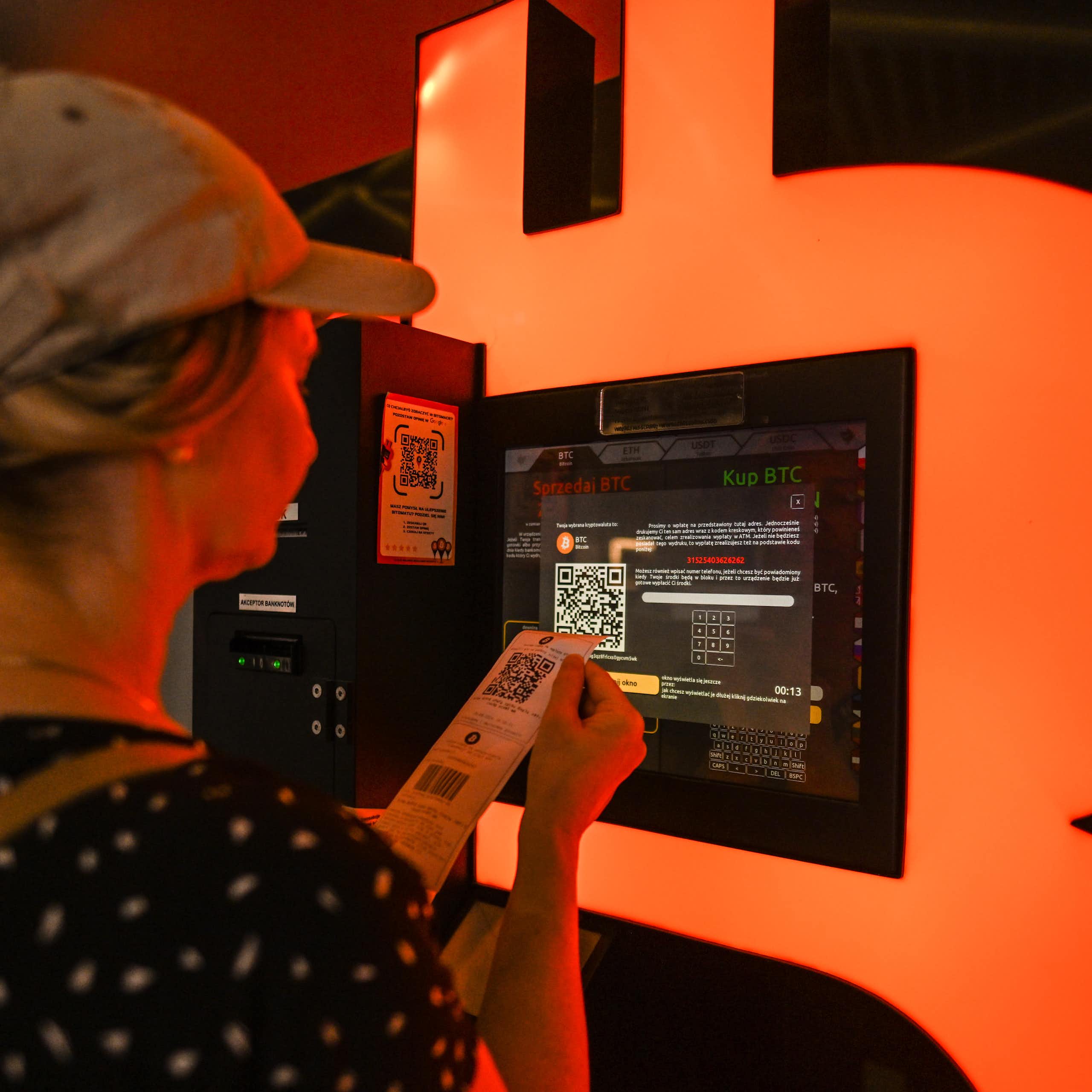 A woman looks intently at a screen while holding a narrow sheet of paper with a QR code on it.