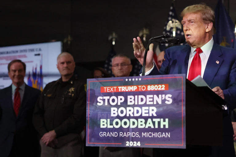 A man in a blue blazer standing at a lectern with a sign that says 'STOP BIDEN'S BORDER BLOODBATH'