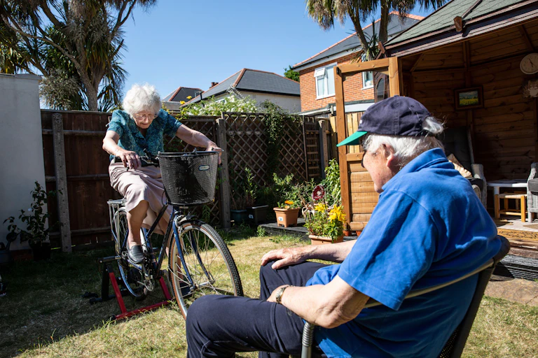An older woman exercising on a bicycle while an older man looks on from the safety of his chair.