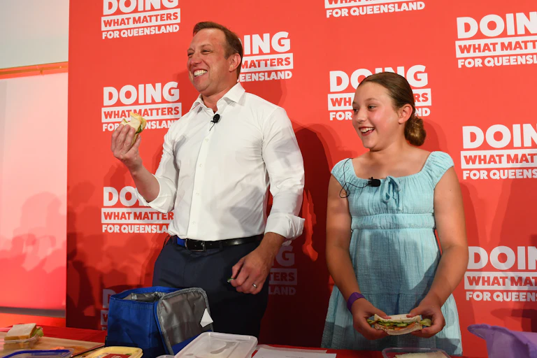 Labor Premier Steven Miles and his daughter Brodie hold sandwiches, standing in front on a Labor campaign poster.