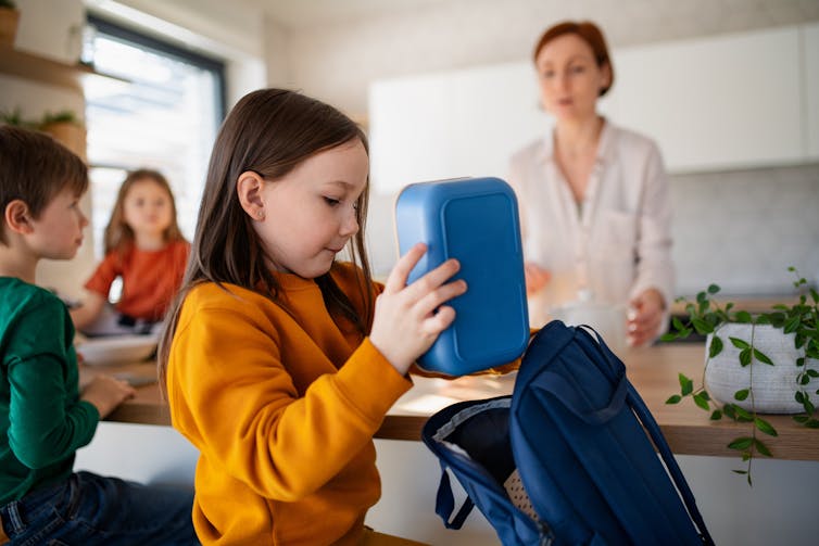 A young girl puts a lunchbox in her bag. A women stands at a kitchen counter behind her.