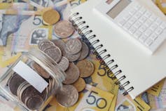 Coins and notes spill from a jar onto a table with a notebook and calculator.
