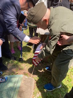 An elderly man in a military uniform bends to plant a small American flag next to a grave in the ground.