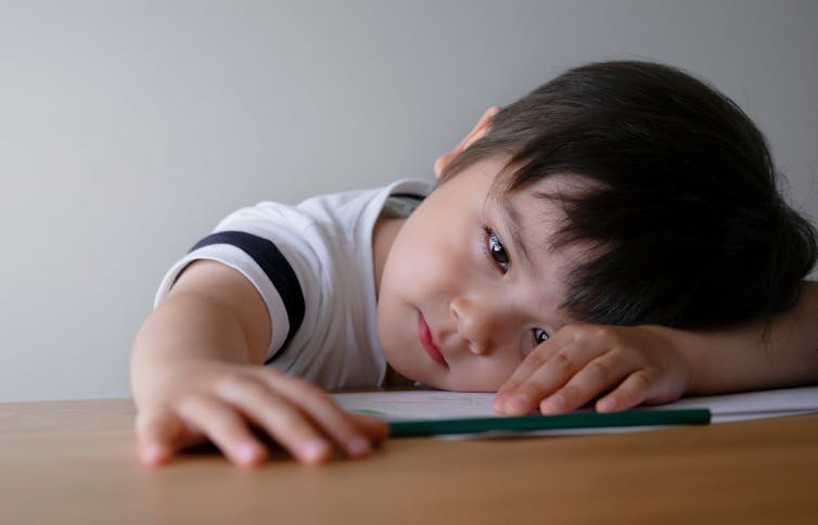 A little boy leans his head against his arm on a table and plays with a pencil.