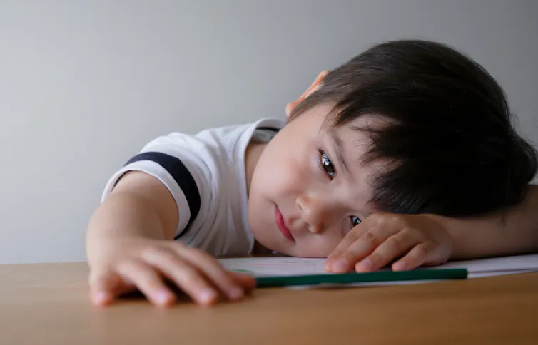 A little boy leans his head against his arm on a table and plays with a pencil.