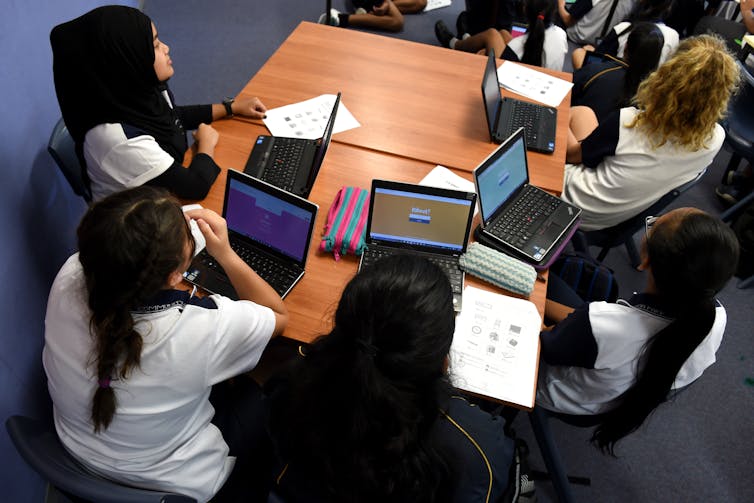Students sit at desks, with laptops in a classroom.