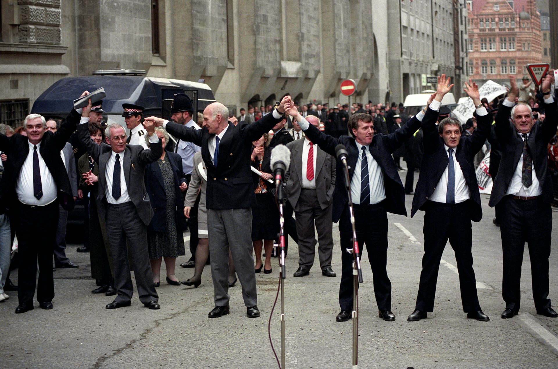 The six Birmingham Six men stand in the street behind microphones with their arms raised.
