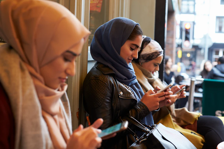 Stock photo of three young women in hijabs sitting outside a cafe and texting