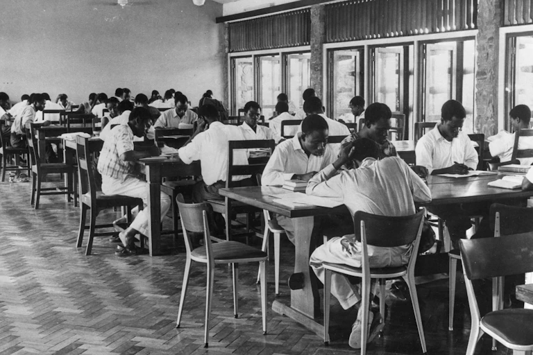A black and white photograph of people sitting at long tables, reading