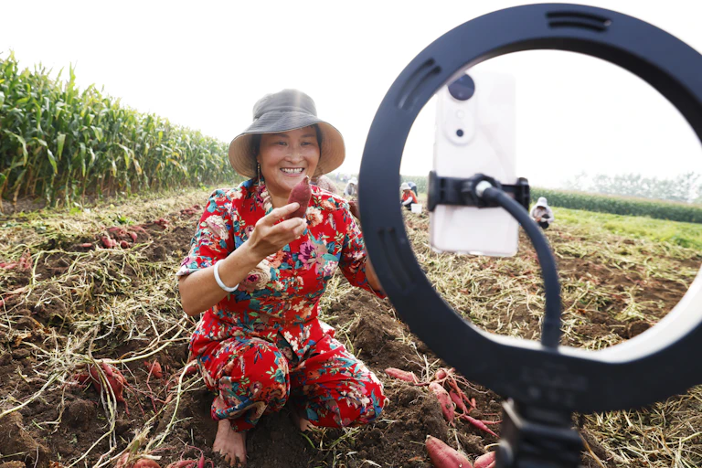 A woman in a hat squats in a field holding a vegetable while being filmed on a phone.