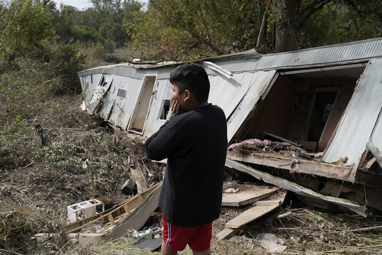 A person puts his hands on his face as he looks at a homes that has been partially crushed, tilted over and strewn with debris.