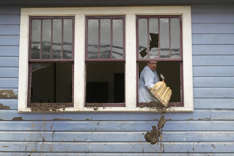 A man scoops mud out a broken window using a snow shovel