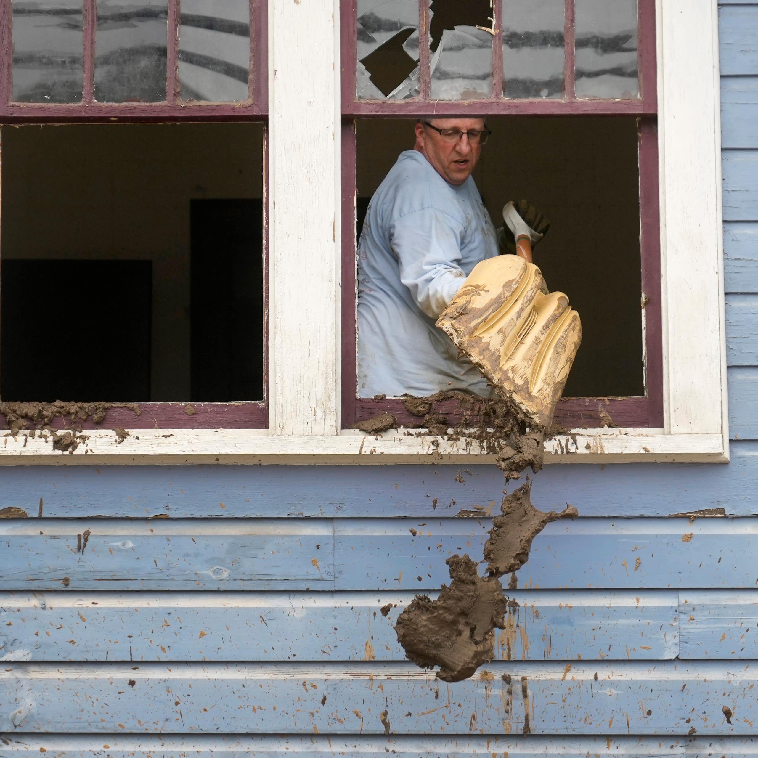 A man scoops mud out a broken window using a snow shovel
