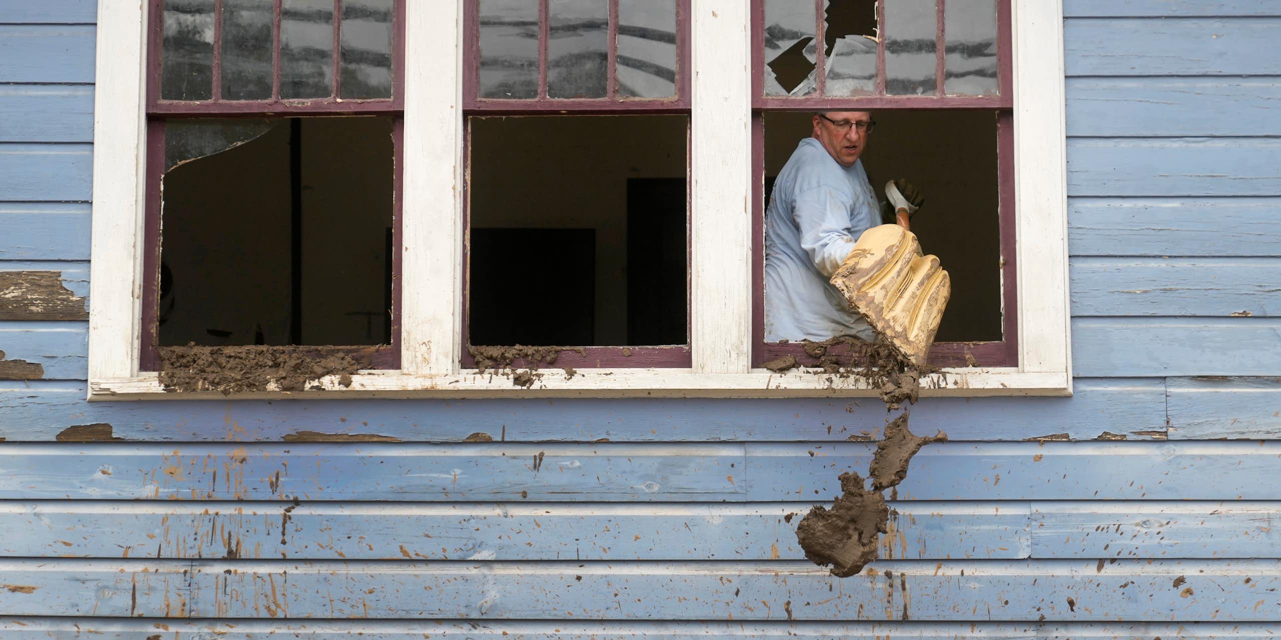A man scoops mud out a broken window using a snow shovel