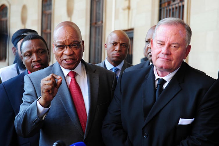 A group of men in suits walks as a balding man in glasses gestures with a fist as he speaks.
