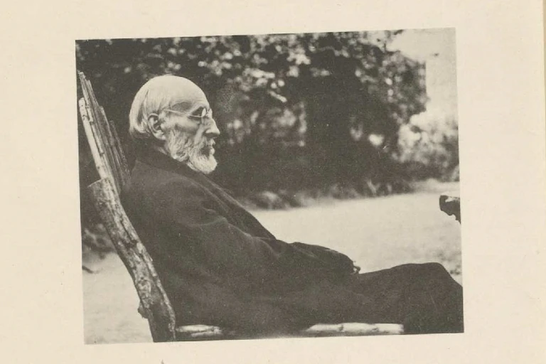Fotografía de un hombre de barba blanca sentado en un jardín de perfil.