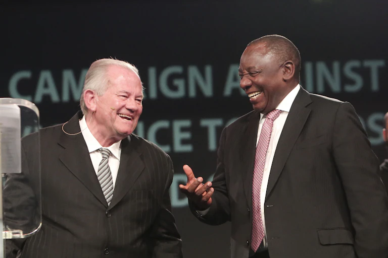 Two men, one caucasians and one African, laugh as they share a joke on a stage, wearing suits and ties.