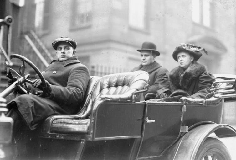 Black and white photo of an old car being driven by a young man. A man and woman sit in the back seats.