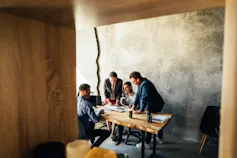 Four colleagues are seen sitting at an office table, drinking coffee and talking.