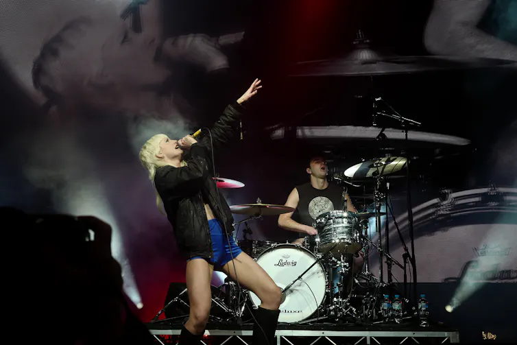 woman hold microphone and sings onstage with drummer playing kit behind her