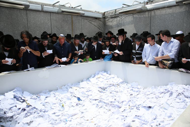 A crowd of men, most of whom wear black jackets, stand around a large white pen filled with pieces of paper.