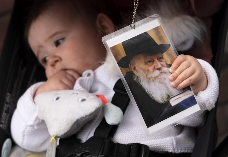 An infant in a white outfit holds onto a laminated photo of an elderly man in a black hat with a long white beard.