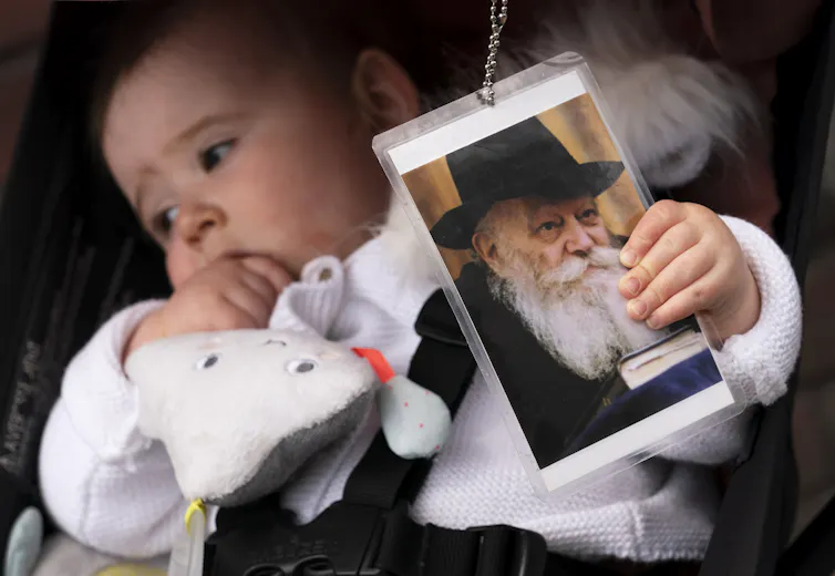 An infant in a white outfit holds onto a laminated photo of an elderly man in a black hat with a long white beard.