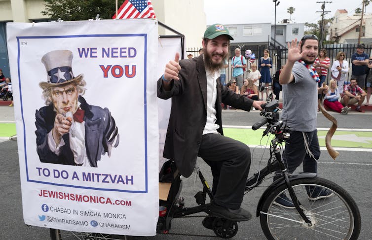 A man makes a thumbs-up sign as he passes by on a bike with an 'Uncle Sam' poster that says 'We need YOU to do a mitzvah.'