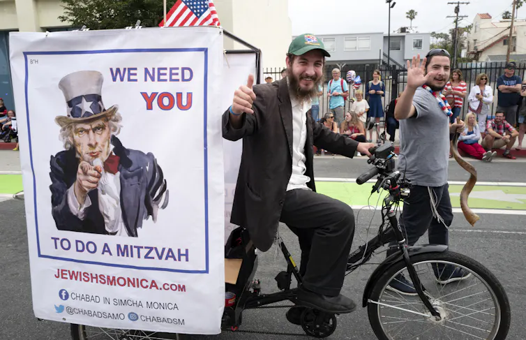 A man makes a thumbs-up sign as he passes by on a bike with an 'Uncle Sam' poster that says 'We need YOU to do a mitzvah.'