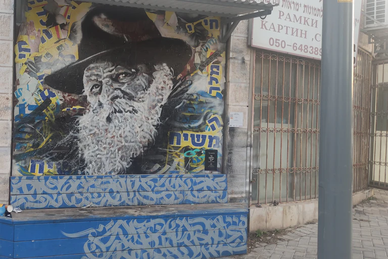 A blue, black and white mural of an elderly man in a black hat, with Hebrew words written in blue and yellow.