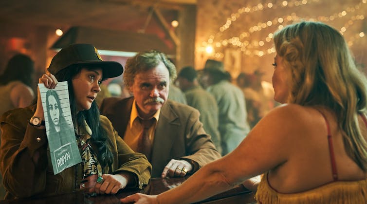 two people hold up a photo of a young girl with dark hair to show a blonde woman behind a bar