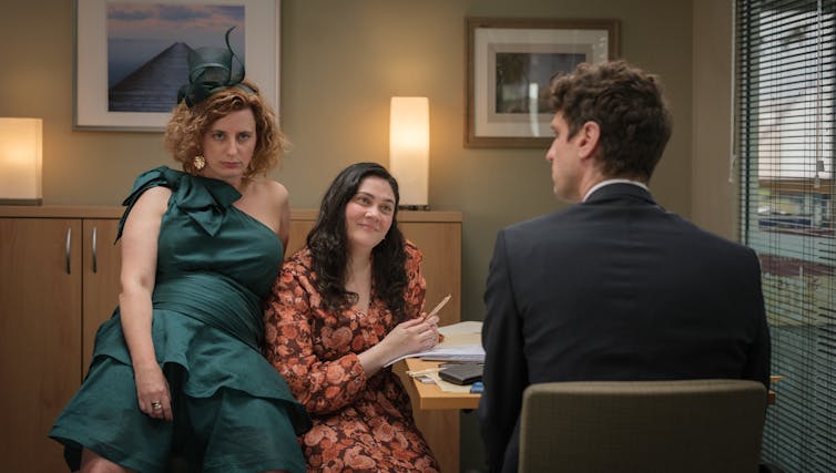 woman in green dress slumps on desk with two other people sitting nearby