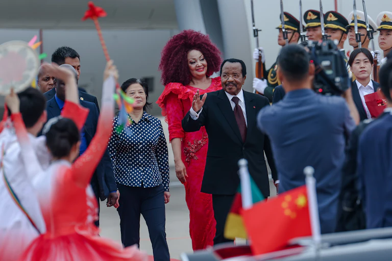A man in suit and tie raises a hand in greeting. He is followed by a woman in a long red dress. A military guard is to one side and a videographer in the foreground.