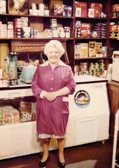An old lady stands in a shop wearing a housecoat.