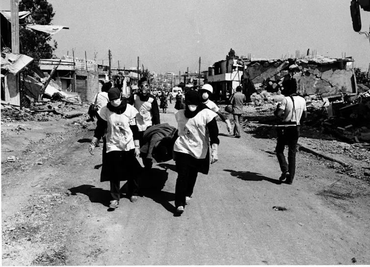 UN relief workers carrying a body on a stretcher through a war-torn city.
