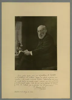 Retrato de un hombre vestido de negro y con barba blanca, sentado ante una mesa con un microscopio.