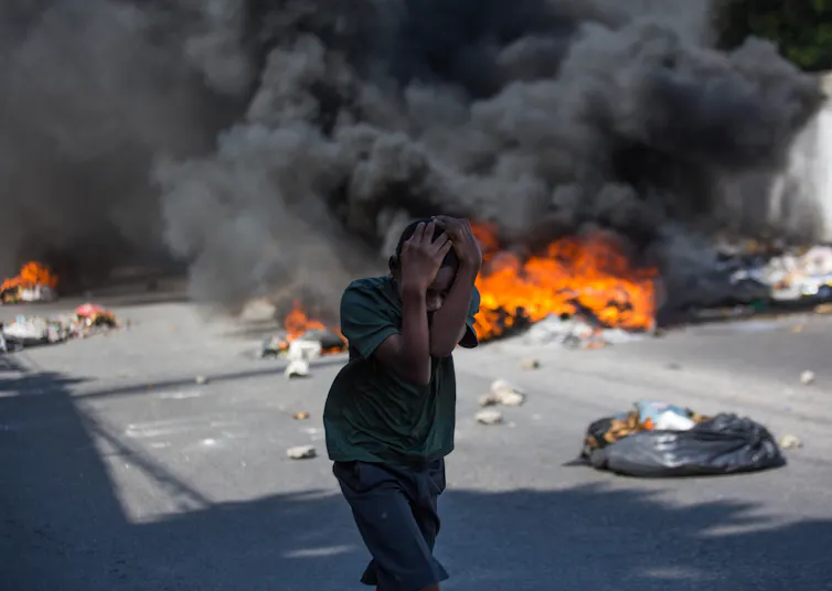 A Haitian child covers his head as a fire rages behind him.